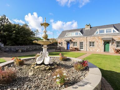 A courtyard with a stone fountain surrounded by flower pots in front of stone cottages with blue and green doors at Tyn Towyn - Ty Glas in Trearddur Bay