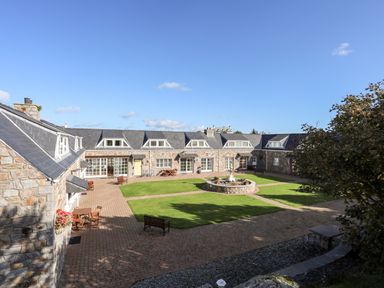 An outdoor courtyard with stone buildings around grass and a central fountain at Tyn Towyn - Ty Glas in Trearddur Bay