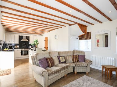 A living room with a beige sofa and patterned pillows next to a kitchen with white cabinets and a black countertop at Tyn Towyn - Ty Glas in Trearddur Bay
