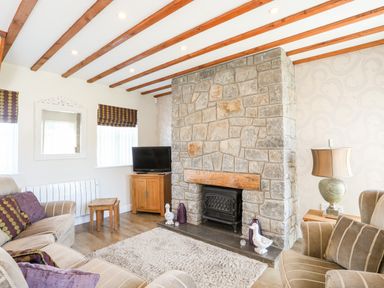 A living room with a stone fireplace armchairs tv and wooden ceiling beams at Tyn Towyn - Ty Glas in Trearddur Bay