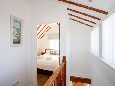 A hallway with wooden railing and ceiling beams looking into a bedroom with a bed and window blinds at Tyn Towyn - Ty Glas in Trearddur Bay