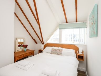 A bedroom with a double bed bedside tables a wooden dresser with a mirror and flowers and slanted wooden ceiling beams at Tyn Towyn - Ty Glas in Trearddur Bay