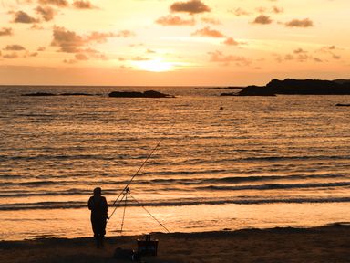A person fishing on a beach at sunset with the sea and rocks in the background at Tyn Towyn - Ty Glas in Trearddur Bay