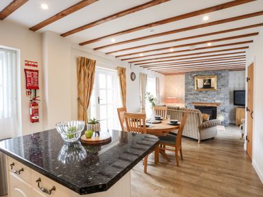 An open living and dining area with wooden beams on the ceiling and a stone fireplace at Tyn Towyn - Ty Oren in Trearddur Bay