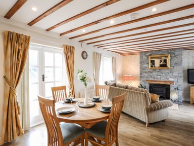 A dining area with wooden table and chairs next to a living room with striped sofas and stone fireplace at Tyn Towyn - Ty Oren in Trearddur Bay