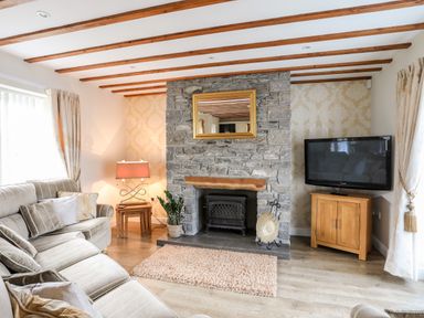A living room with a stone fireplace wooden beams a sofa and a television at Tyn Towyn - Ty Oren in Trearddur Bay