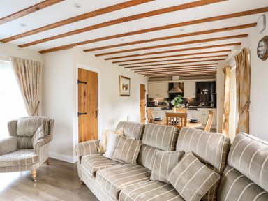 An open living room and kitchen with striped sofa and armchair wooden beams on ceiling and dining table with chairs at Tyn Towyn - Ty Oren in Trearddur Bay