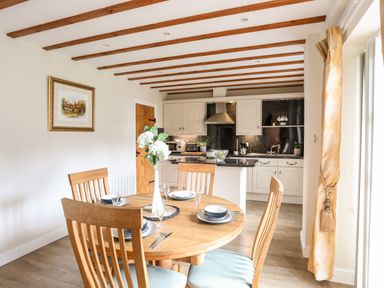 A dining area with a round wooden table set with dishes and glasses next to a kitchen with white cabinets and wooden ceiling beams at Tyn Towyn - Ty Oren in Trearddur Bay