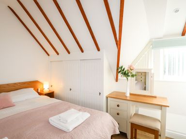 A bedroom with a bed pink bedspread a bedside table lamp a dressing table with a mirror and flowers at Tyn Towyn - Ty Oren in Trearddur Bay