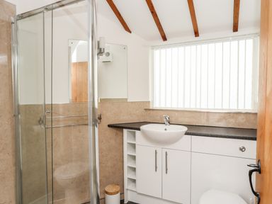 A bathroom with a glass shower, a white sink and cabinet, and a window with vertical blinds at Tyn Towyn - Ty Oren in Trearddur Bay