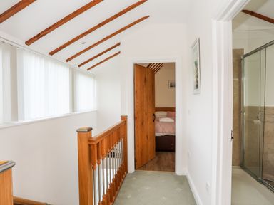 A hallway with wooden railing and ceiling beams leading to a bedroom and a bathroom with a glass shower door at Tyn Towyn - Ty Oren in Trearddur Bay