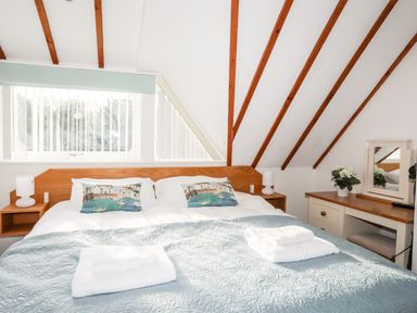 A bedroom with a double bed with decorative pillows and folded towels a dressing table with a mirror and plant and slanted ceiling with wooden beams at Tyn Towyn - Ty Oren in Trearddur Bay