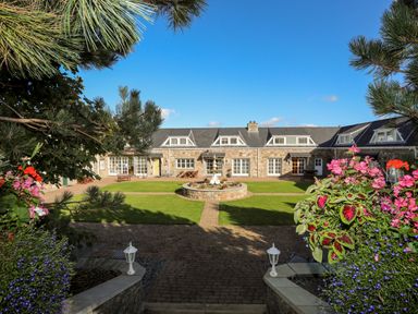 A stone building with dormer windows and a central fountain in a courtyard with grass and flowers at Tyn Towyn - Ty Oren in Trearddur Bay