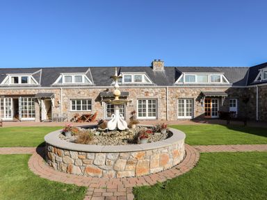 A stone building with multiple dormer windows and white framed doors surrounding a central stone fountain with plants in a garden at Tyn Towyn - Ty Oren in Trearddur Bay