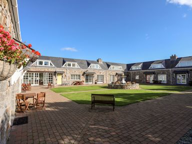 A courtyard with stone buildings around a grass area and outdoor furniture at Tyn Towyn - Ty Oren in Trearddur Bay