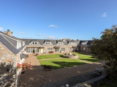 An outdoor courtyard with grassy areas and a central fountain surrounded by stone buildings at Tyn Towyn - Ty Oren in Trearddur Bay