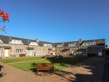 A stone courtyard with grass, a fountain, benches, and attached houses at Tyn Towyn - Ty Oren in Trearddur Bay