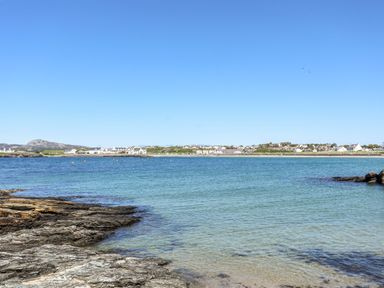 A coastal scene with rocky shore in the foreground and houses across the water at Tyn Towyn - Ty Oren in Trearddur Bay