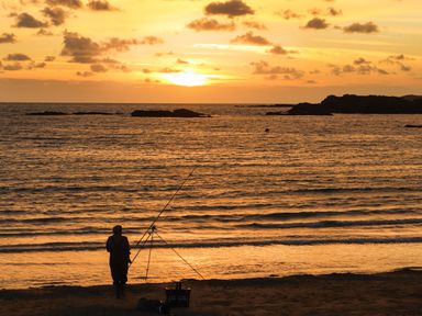 A person fishing on the beach during sunset with the sea and rocks in the background at Tyn Towyn - Ty Oren in Trearddur Bay