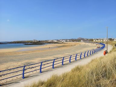 A sandy beach with a blue railing and pathway near houses at Tyn Towyn - Ty Oren in Trearddur Bay