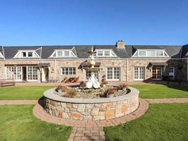 A stone building with multiple windows and a courtyard featuring a circular stone fountain with swan statues in front of it at Tyn Towyn - Ty Piws in Trearddur Bay