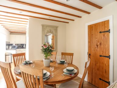 A dining room with a round wooden table set with bowls plates and glasses a potted plant in the center wooden chairs a wooden door and a large decorative wall mirror at Tyn Towyn - Ty Piws in Trearddur Bay