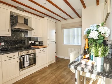 A kitchen with white cabinets a built-in oven a toaster and a small dining area with flowers and a bottle at Tyn Towyn - Ty Piws in Trearddur Bay