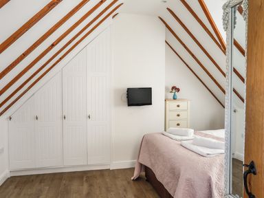 A bedroom with a bed covered with a pink quilt and white towels a chest of drawers with a vase of flowers a wall-mounted television and built in cupboards under a slanted ceiling at Tyn Towyn - Ty Piws in Trearddur Bay