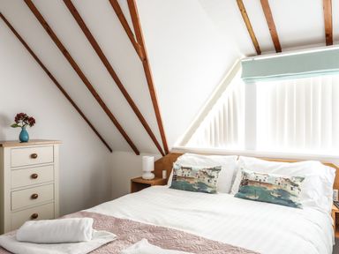 A bedroom with a bed white linens wooden beams on the ceiling a chest of drawers with a vase and a nightstand with a lamp at Tyn Towyn - Ty Piws in Trearddur Bay
