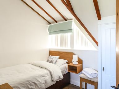 A bedroom with a single bed a pillow a bedside table with a lamp folded towels on a stool under a slanted ceiling with wooden beams at Tyn Towyn - Ty Piws in Trearddur Bay