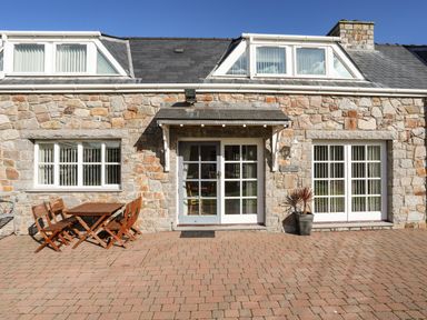 A stone house exterior with white-framed windows and doors and a wooden table with chairs on a paved patio at Tyn Towyn - Ty Piws in Trearddur Bay