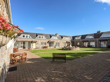 An outdoor courtyard with stone buildings surrounding a grass area and outdoor seating at Tyn Towyn - Ty Piws in Trearddur Bay