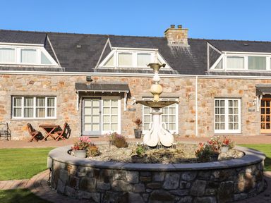 A stone building with glass doors and windows a fountain with swan sculptures in front and flower pots on the fountain at Tyn Towyn - Ty Piws in Trearddur Bay
