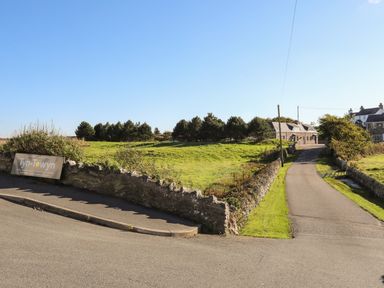 A paved road leading to stone buildings with a low stone wall and grassy fields on each side at Tyn Towyn - Ty Piws in Trearddur Bay