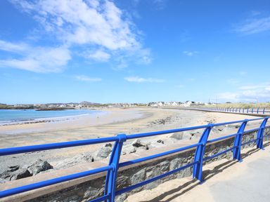 A beach with a blue railing along a stone wall and houses in the distance at Tyn Towyn - Ty Piws in Trearddur Bay
