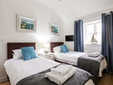 A bedroom with two single beds blue cushions bedside lamp and window with blue curtains at Tyn Towyn - The Stables in Trearddur Bay