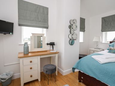 A bedroom with a white desk, round mirror, stool, wall decor, bed with blue bedding, and windows with gray blinds at Tyn Towyn - The Stables in Trearddur Bay