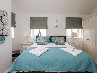 A bedroom with a bed covered in blue bedspread and pillows between two windows with grey blinds at Tyn Towyn - The Stables in Trearddur Bay