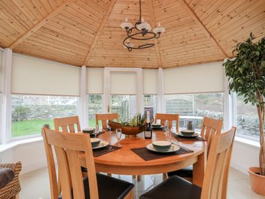 A dining area with a round wooden table set with plates glasses and a bottle under a wooden ceiling with windows and blinds at Tyn Towyn - The Stables in Trearddur Bay