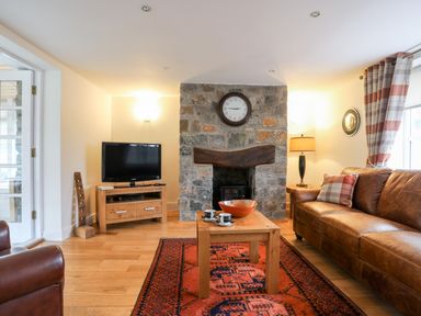 A living room with a leather sofa a stone fireplace a wooden coffee table and a television at Tyn Towyn - The Stables in Trearddur Bay