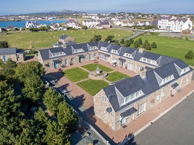 A courtyard with a fountain surrounded by stone buildings with dormer windows and a view of houses and fields at Tyn Towyn - The Stables in Trearddur Bay