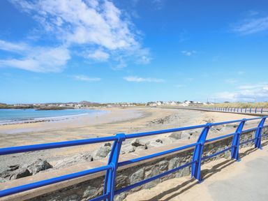 A sandy beach with a blue railing along a paved walkway and houses in the background at Tyn Towyn - The Stables in Trearddur Bay