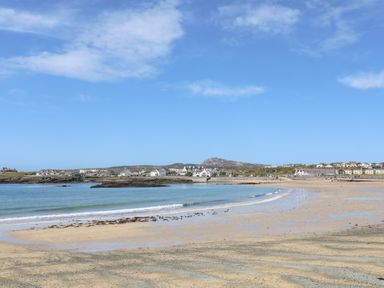 A sandy beach with gentle waves and scattered seaweed with a coastal village and hills in the background at Tyn Towyn - The Stables in Trearddur Bay