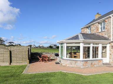 A stone house with a sunroom overlooking a paved patio with wooden outdoor furniture and a barbecue grill at Tyn Towyn - The Stables in Trearddur Bay
