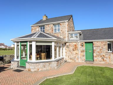 A stone house with green doors and large windows and a paved walkway with grass at Tyn Towyn - The Stables in Trearddur Bay