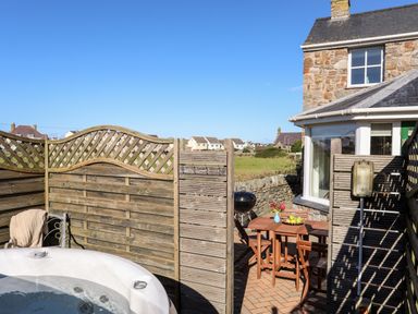 An outdoor patio area with a wooden table and chairs a hot tub and a stone house at Tyn Towyn - The Stables in Trearddur Bay