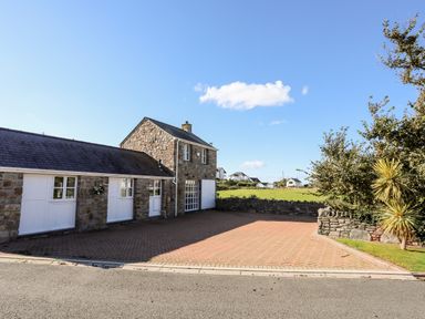 A stone building with white doors and windows next to a paved driveway and trees at Tyn Towyn - The Stables in Trearddur Bay