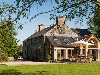 A stone house with outdoor wooden table and chairs under an umbrella in a garden at Tyddyn Morthwyl in Criccieth