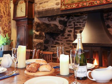 A dining table set with bread rolls champagne bottle candles and glasses in front of a stone fireplace at Tyddyn Morthwyl Criccieth