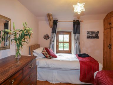 A bedroom with a single bed red bedspread wooden dresser with flowers and a window with grey curtains at Tyddyn Morthwyl in Criccieth